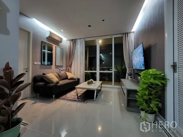 Condominium for rent in Huai Khwang, Bangkok - Cozy living area at night with sofa, wall-mounted TV and floor-to-ceiling windows.