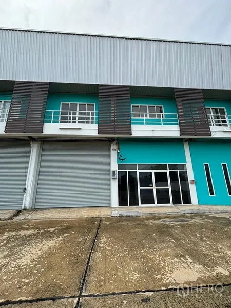 Warehouse for rent in Phra Samut Chedi, Samut Prakan - Teal warehouse exterior with twin roller shutters, balcony windows and a glass-front office entrance.