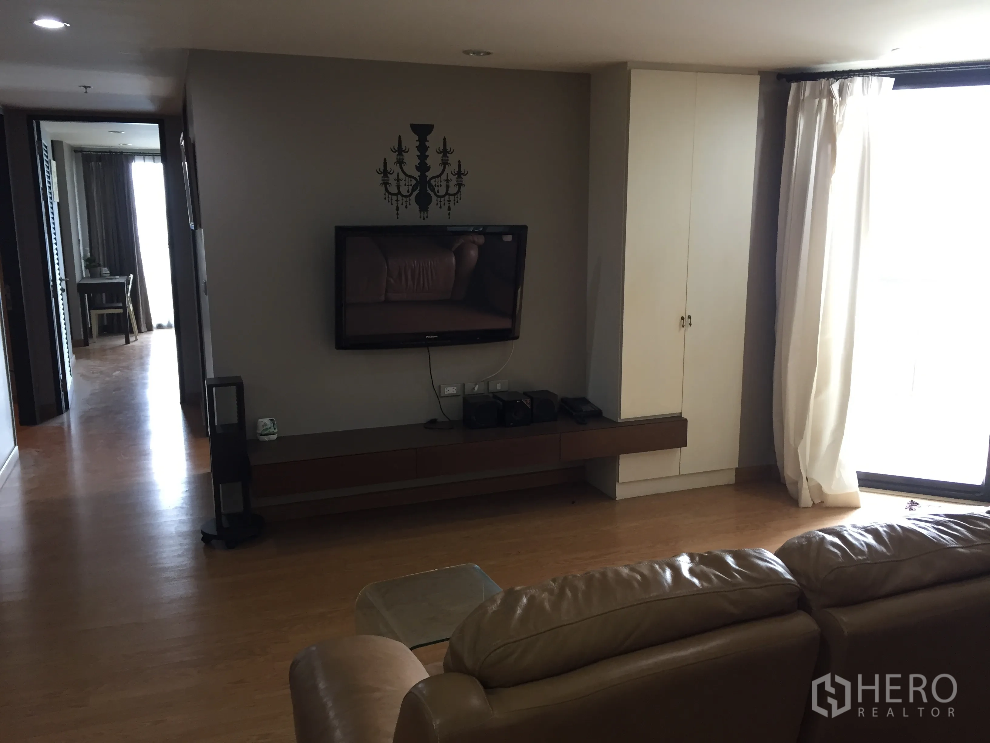 Condominium for rent in Watthana, Bangkok - Living area with wall-mounted TV, long media console, floor-to-ceiling window with curtains, and warm wood flooring.