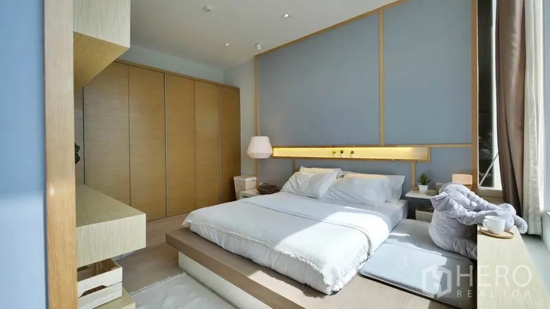 Condominium for rent or sale in Khlong Toei, Bangkok - Sunlit bedroom with platform bed, large wardrobes and a soft blue feature wall.
