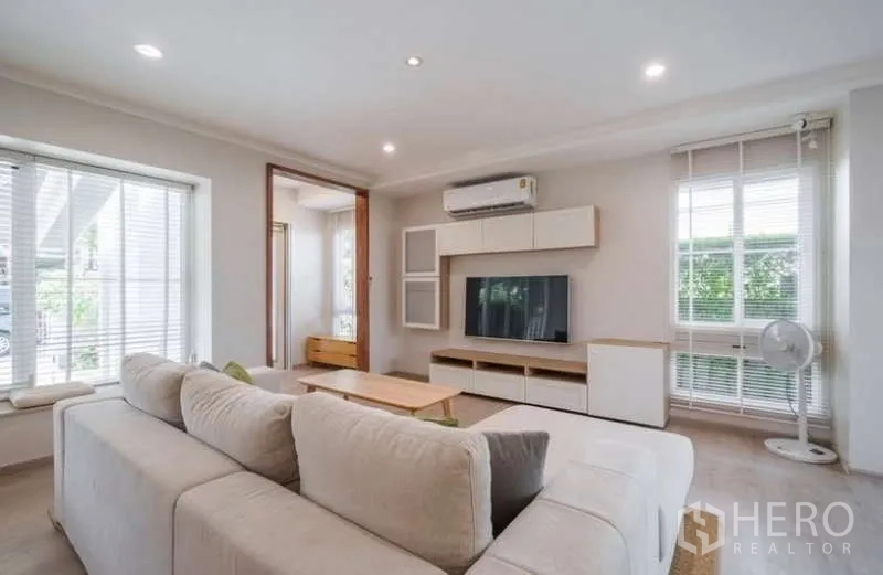 Detached House for rent in Prawet, Bangkok - Living room with L-shaped sofa, media wall, air conditioner and bright windows.