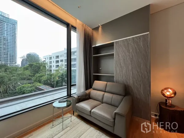 Condominium for rent in Pathum Wan, Bangkok - Bright living area with leather sofa, built-in shelving and floor-to-ceiling window overlooking treetops and city.