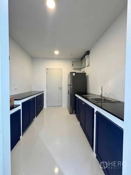 Detached House for rent or sale in Bang Phli, Samut Prakan - Galley kitchen with dual black countertops, navy cabinetry, sink and fridge in a bright white room.