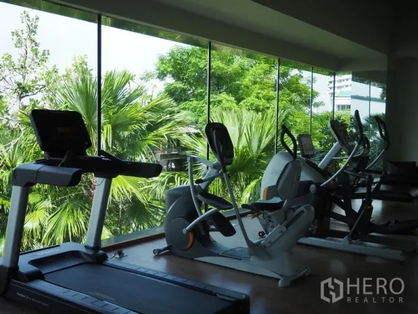 Condominium for rent or sale in Bangkok - Condominium fitness room with treadmills and ellipticals facing lush greenery through floor-to-ceiling windows.
