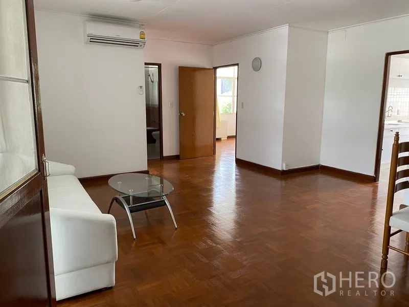 Apartment for rent in Khlong Toei, Bangkok - Living room with white sofa, glass table, split air-conditioning and doorways to other rooms.