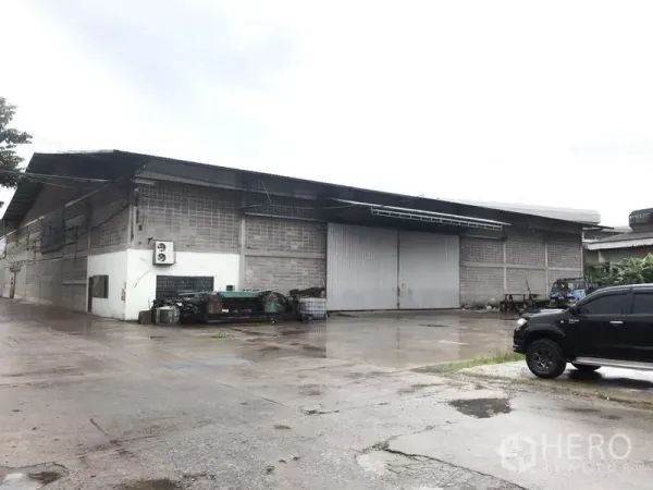 货栈 出租在Mueang Pathum Thani, Pathum Thani - 仓库外观，配有大型金属推拉门与宽阔混凝土堆场，雨天环境。
