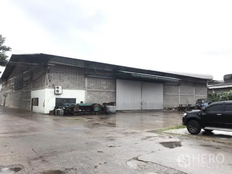 货栈 出租在Mueang Pathum Thani, Pathum Thani - 仓库外观，配有大型金属推拉门与宽阔混凝土堆场，雨天环境。