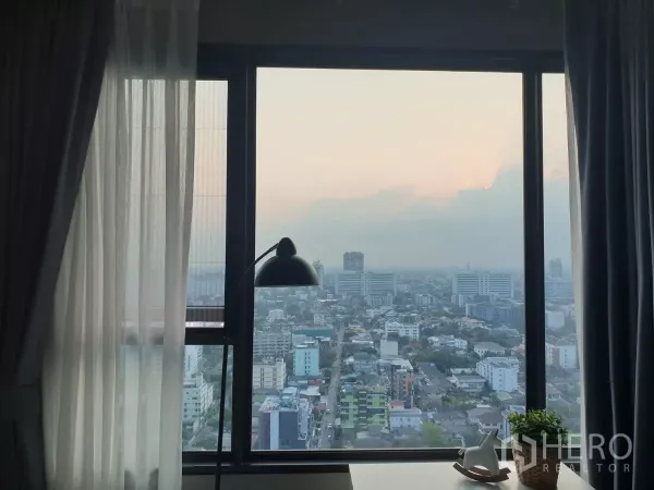 คอนโด ให้เช่าในChatuchak, Bangkok - วิวเมืองจตุจักรจากหน้าต่างชั้นสูงยามพระอาทิตย์ตก พร้อมม่านและโคมไฟตั้งโต๊ะ