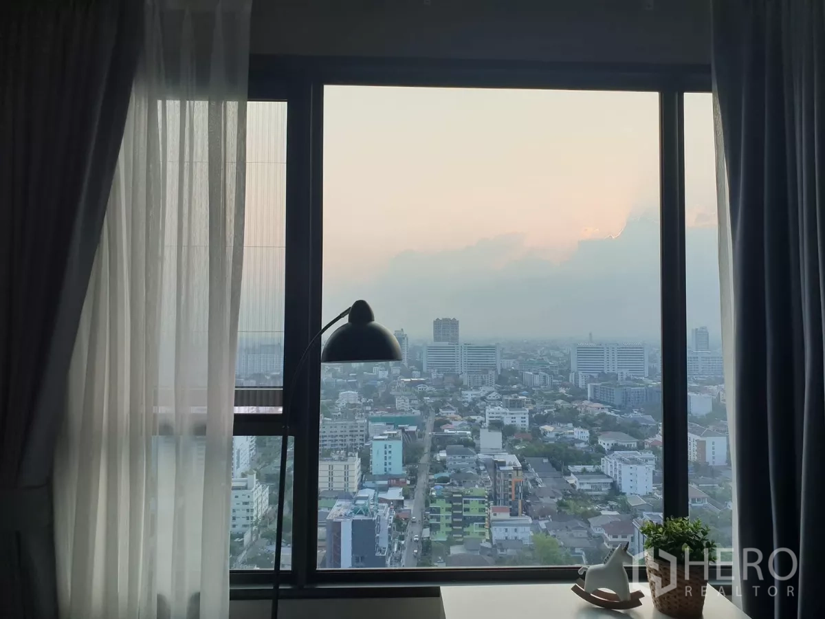 คอนโด ให้เช่าในChatuchak, Bangkok - วิวเมืองจตุจักรจากหน้าต่างชั้นสูงยามพระอาทิตย์ตก พร้อมม่านและโคมไฟตั้งโต๊ะ