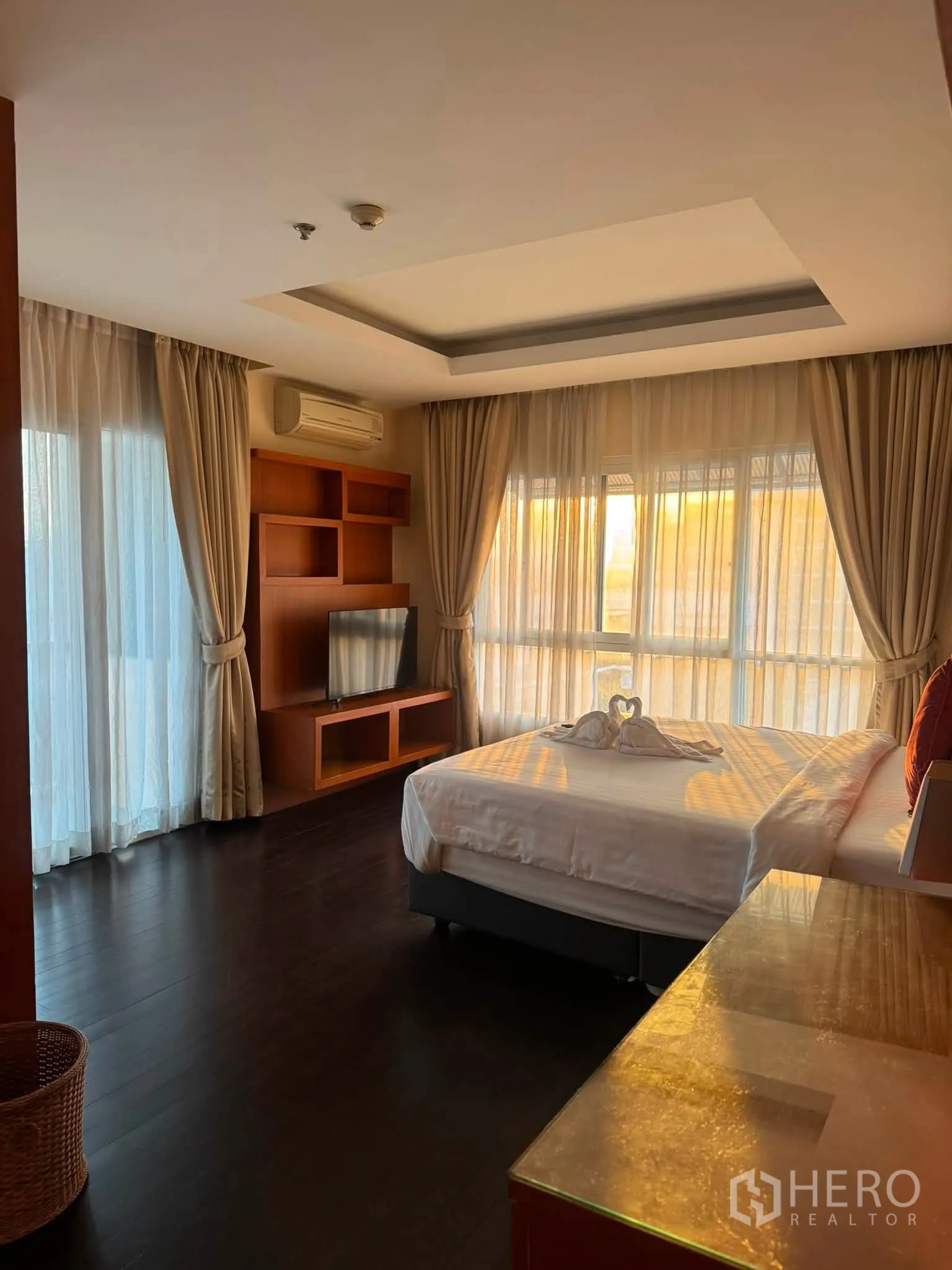 Condominium for rent in Khlong Toei, Bangkok - Sunlit bedroom with large windows, sheer curtains, built-in wood TV unit, and dark wood flooring.