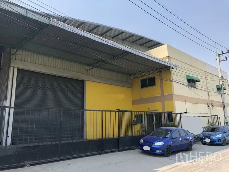 Factory for rent in Pathum Thani - Street view of the factory facade with metal awning, tall black gate and roadside parking in an industrial area.