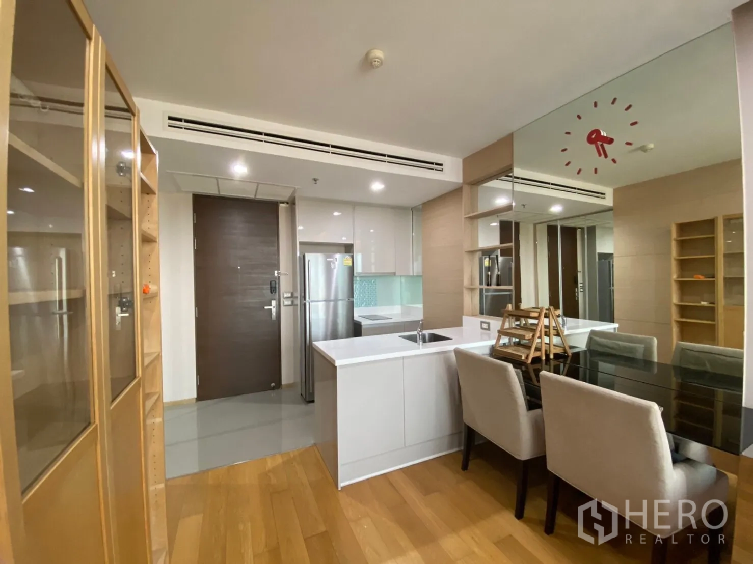 Condominium for rent in Ratchathewi, Bangkok - Dining nook with glass-top table and upholstered chairs beside a sleek white kitchenette and mirrored wall.