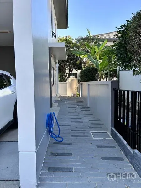 Detached House for rent or sale in Prawet, Bangkok - Side walkway with tiled path, garden plants, and a water tank.