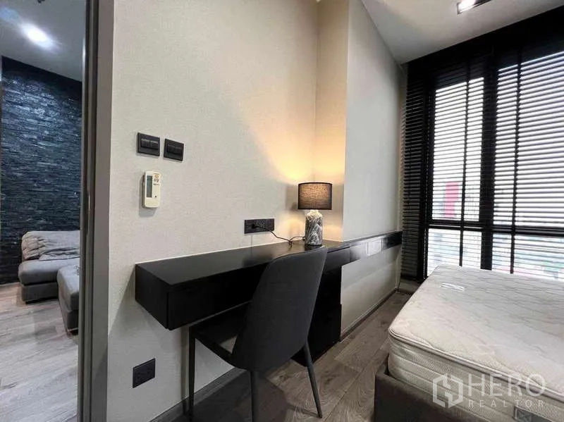 Condominium for rent - Bedroom study corner with a sleek desk and lamp beside large windows with blinds; living room visible beyond.