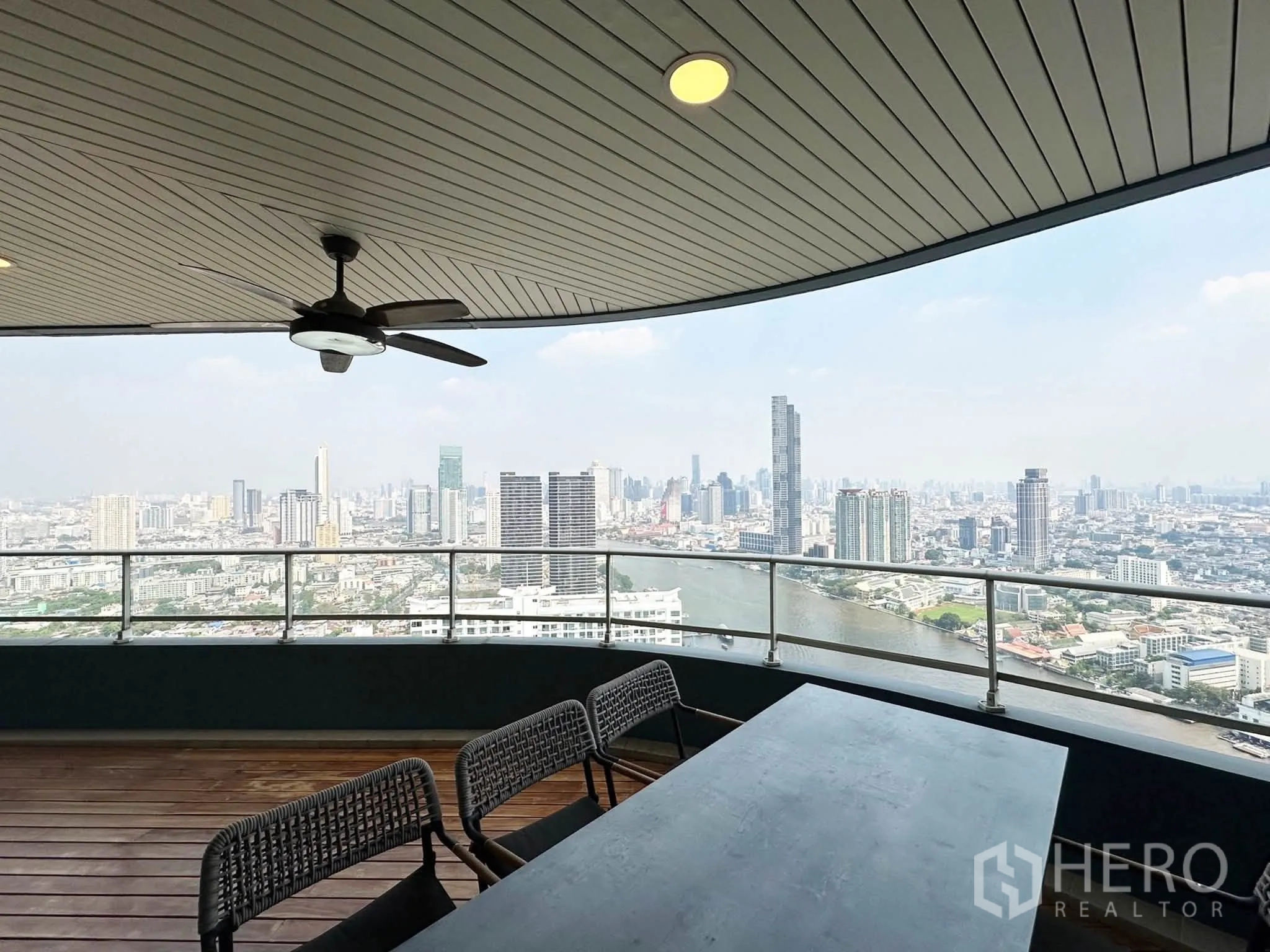 Condominium for sale in Khlong San, Bangkok - Covered balcony with ceiling fan and long table facing a panoramic city and river view.