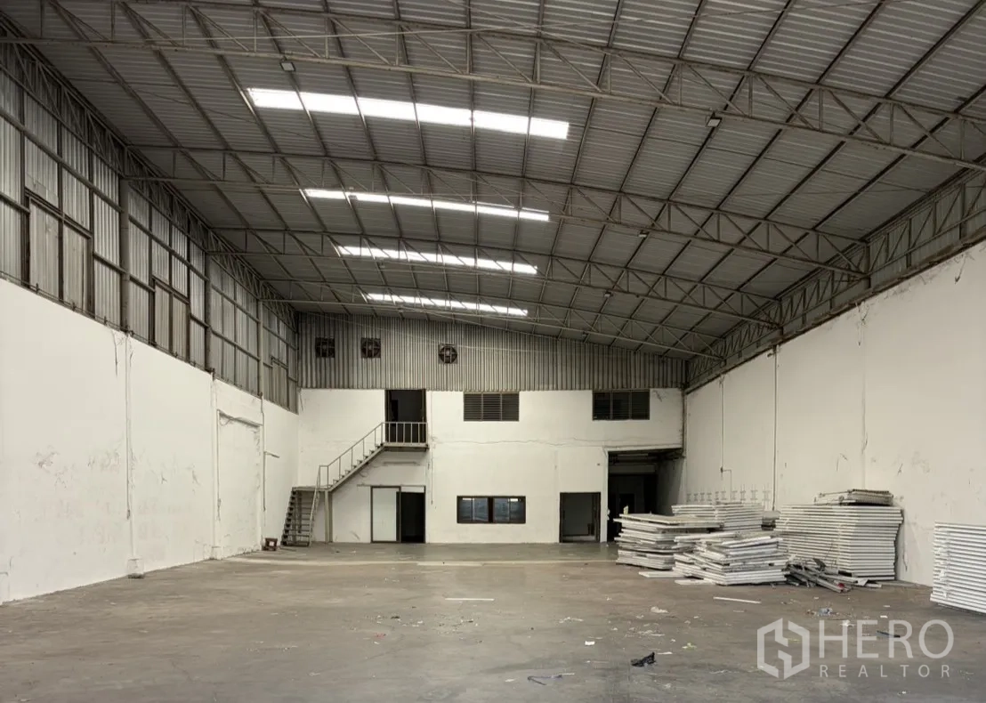 仓库 for rent in Khlong Luang, Pathum Thani - 仓库内部空间宽敞，钢结构高屋顶与采光板，混凝土地面，带有楼梯与窗户的夹层办公室，侧面堆放材料。