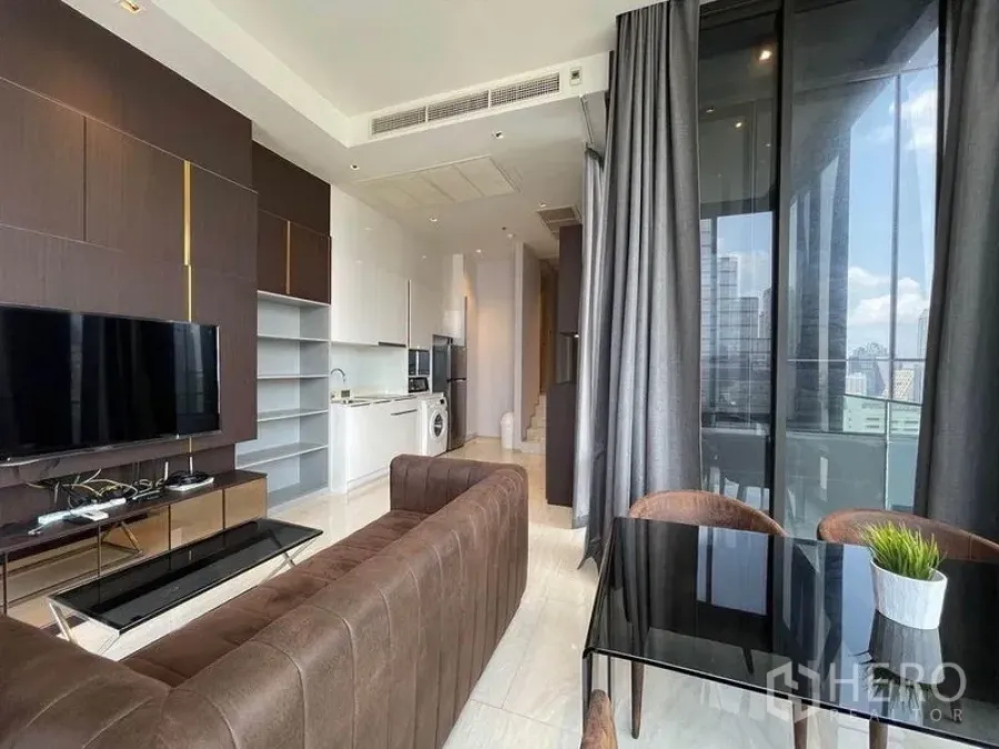 Condominium for rent in Bang Rak, Bangkok - Open living, dining and kitchen layout with brown sofa, TV wall and corner glass windows showing a city view.