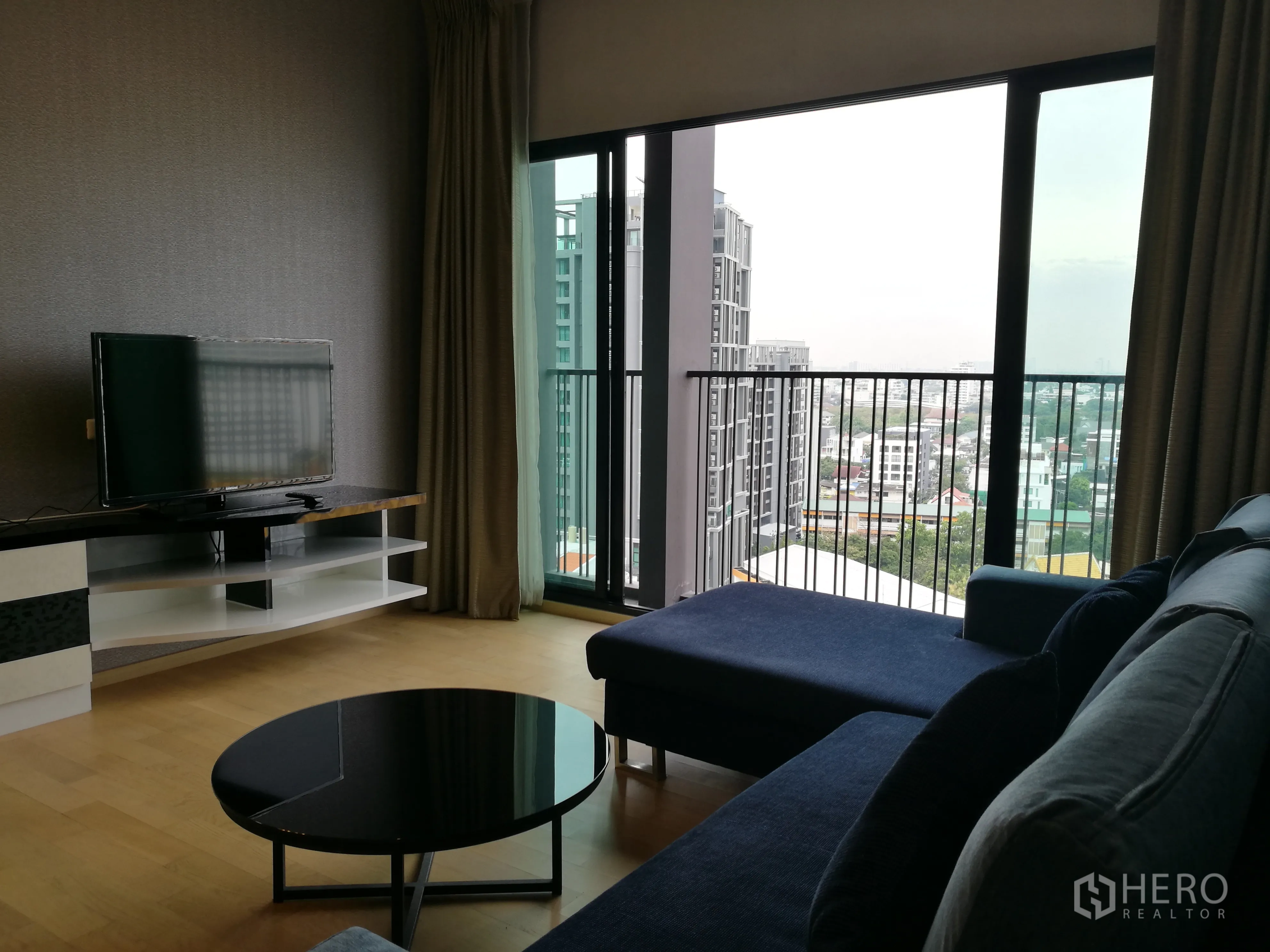 Condominium for rent in Watthana, Bangkok - Living room with blue sectional sofa, TV console, and floor‑to‑ceiling sliding doors opening to a balcony overlooking the city.