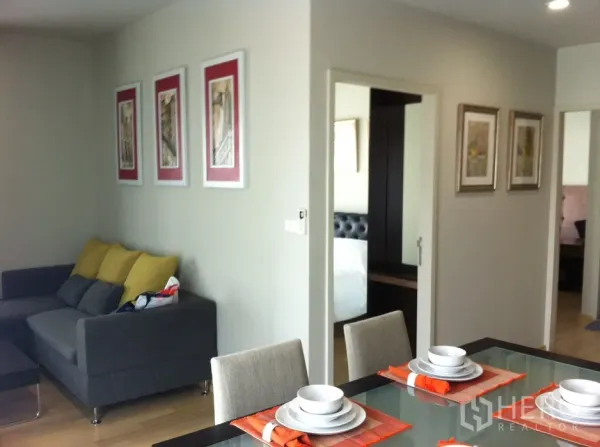 คอนโด ให้เช่าในBangkok - มุมรับประทานอาหารโต๊ะกระจกพร้อมชุดจาน ถัดไปเป็นโซฟาสีเทาพนักพิงเหลือง และทางเข้าห้องนอนที่มีกระจกตู้เสื้อผ้า