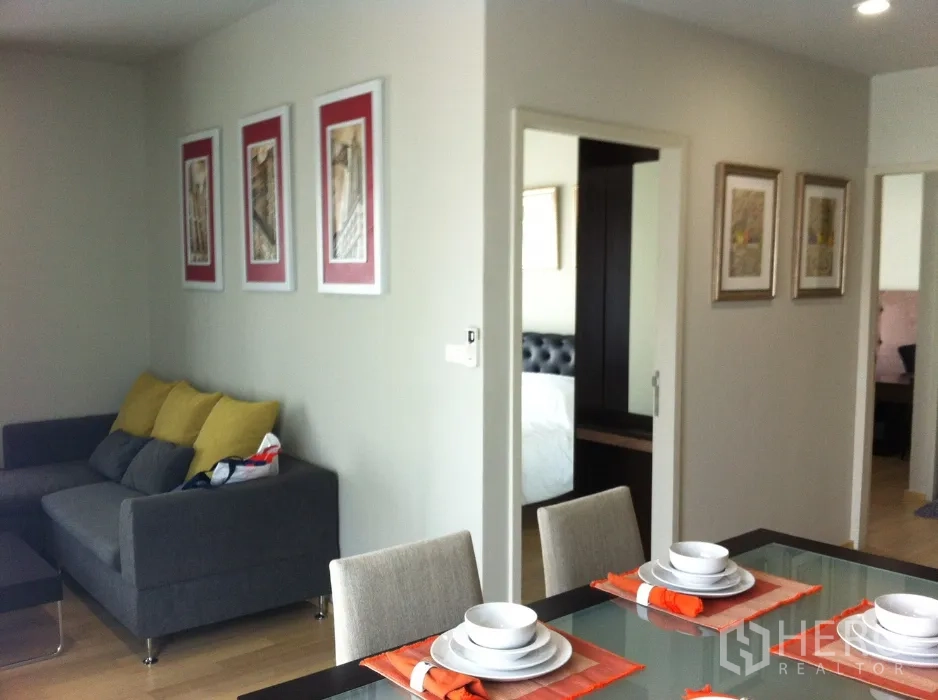 คอนโด ให้เช่าในBangkok - มุมรับประทานอาหารโต๊ะกระจกพร้อมชุดจาน ถัดไปเป็นโซฟาสีเทาพนักพิงเหลือง และทางเข้าห้องนอนที่มีกระจกตู้เสื้อผ้า