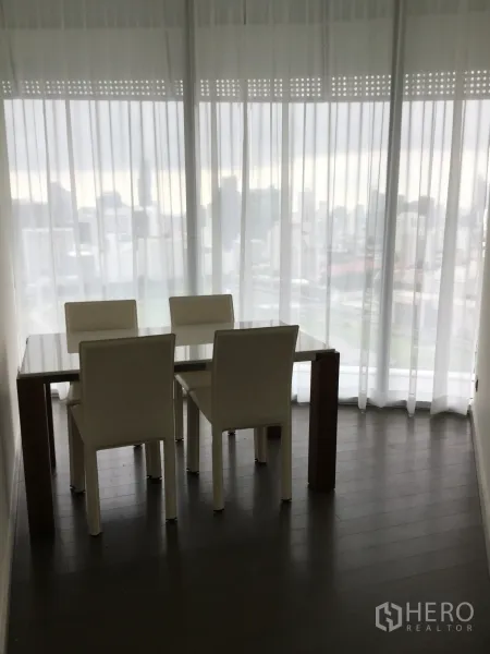 คอนโด ให้เช่าในPathum Wan, Bangkok - มุมทานอาหารพร้อมเก้าอี้สี่ตัวและโต๊ะกระจก ข้างหน้าต่างสูงจรดเพดานเห็นวิวเมือง.