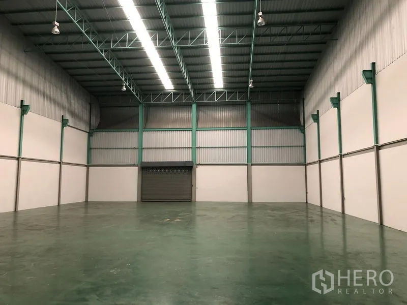 Factory for rent in Samut Prakan - High‑ceiling warehouse bay with steel trusses, LED lighting and a roller shutter door.