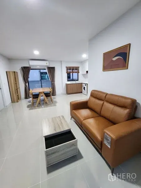 Townhouse for rent in Bang Phli, Samut Prakan - Open-plan living, dining, and kitchen area with a leather sofa, large windows, and tiled floors.