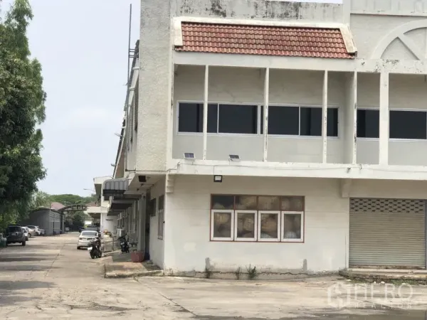Warehouse for rent in Bang Kapi, Bangkok - Close view of the warehouse corner with shaded walkway, windows and a roller shutter beside a tree-lined driveway.