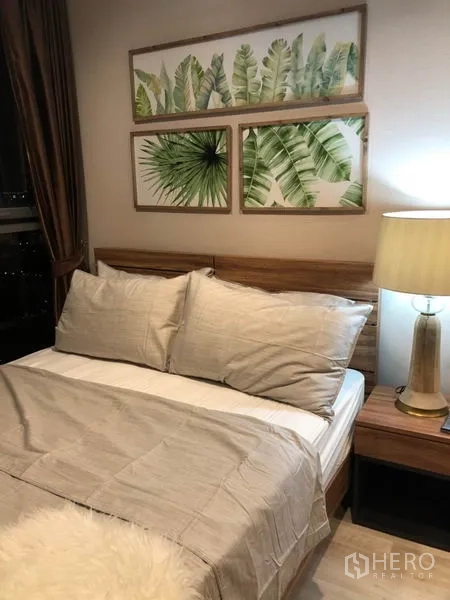 Condominium for rent or sale in Phra Khanong, Bangkok - Bedroom corner with wooden bed, bedside lamp and tropical leaf artwork.