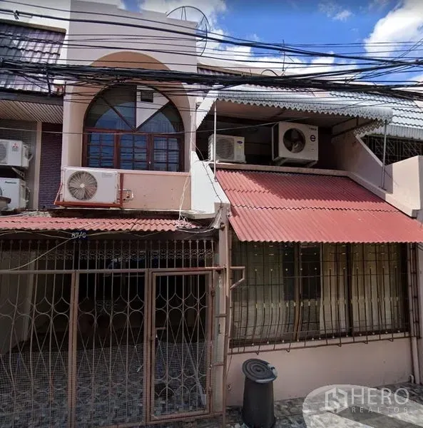 ทาวน์เฮาส์ ให้เช่าในKhlong Toei, Bangkok - หน้าบ้านทาวน์เฮ้าส์สองชั้น มีประตูรั้ว กันสาดโลหะ และคอมเพรสเซอร์แอร์ด้านนอก