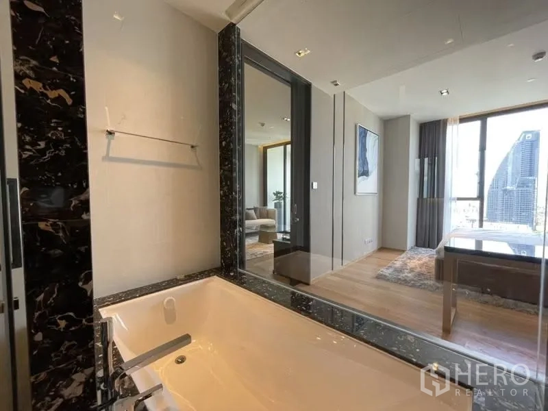 Condominium for rent or sale in Khlong Toei, Bangkok - Modern bathroom with black marble-trimmed bathtub and a glass wall looking toward the bedroom and city view.