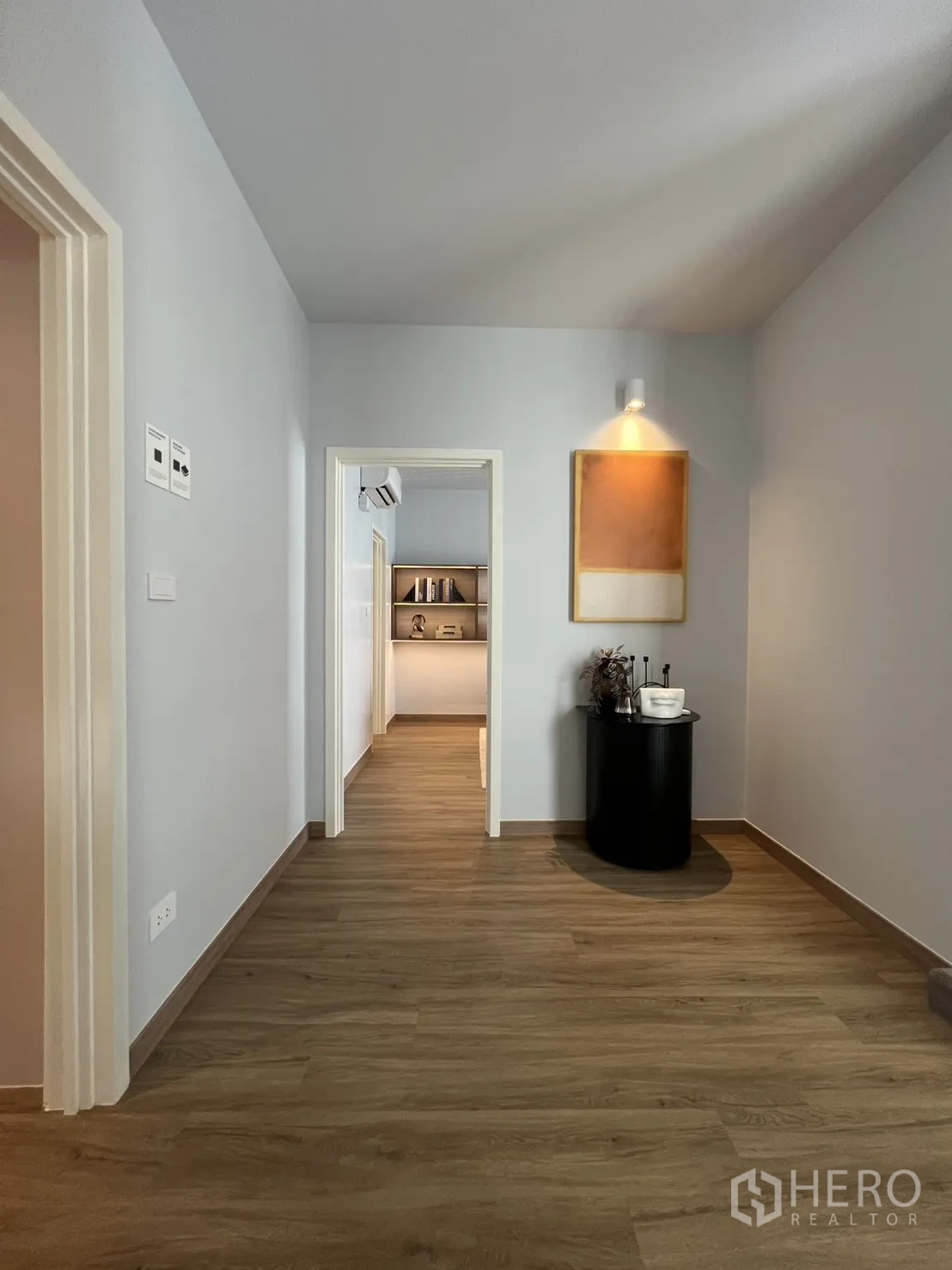 Townhouse for rent in Mueang Samut Prakan, Samut Prakan - Bright hallway with wood flooring, niche wall light, and view into a study with shelving.