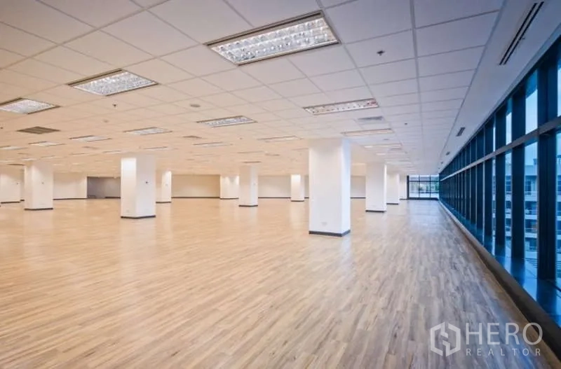 办公空间 出租在Phaya Thai, Bangkok - 转角办公空间，浅木地板、白色立柱、吊顶照明，一侧为整排大面积玻璃。