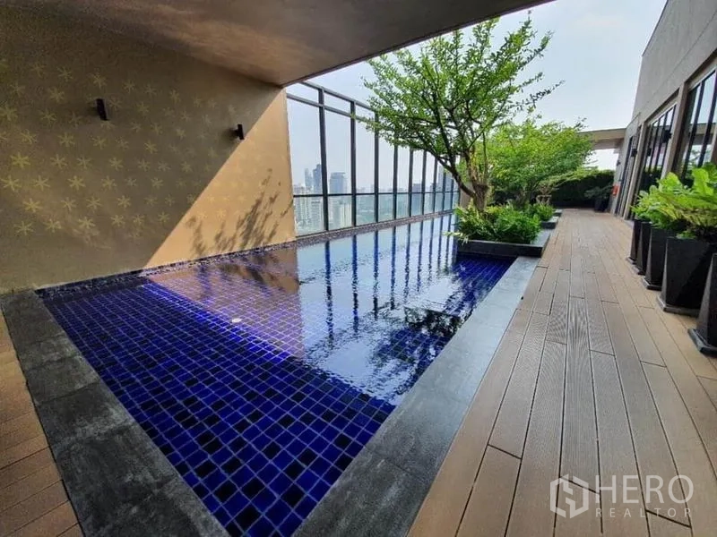 Condominium for sale in Ratchathewi, Bangkok - Rooftop swimming pool with blue mosaic tiles, deck seating and skyline views beside planters and trees.
