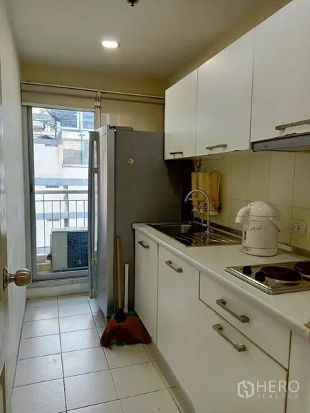 Condominium for rent in Bang Rak, Bangkok - Galley kitchen with white cabinets, sink, electric cooktop, fridge and balcony access.