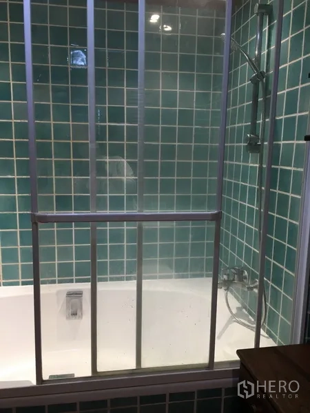 Condominium for rent in Pathum Wan, Bangkok - Bathtub with glass shower screen set against green tiled wall.