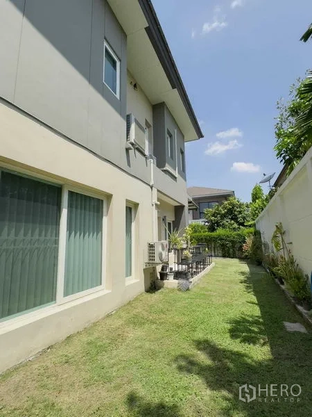 บ้าน ให้เช่าหรือขายในBang Khen, Bangkok - สนามหญ้าด้านข้างและผนังภายนอกบ้านสองชั้น มีหน้าต่างและคอมเพรสเซอร์แอร์