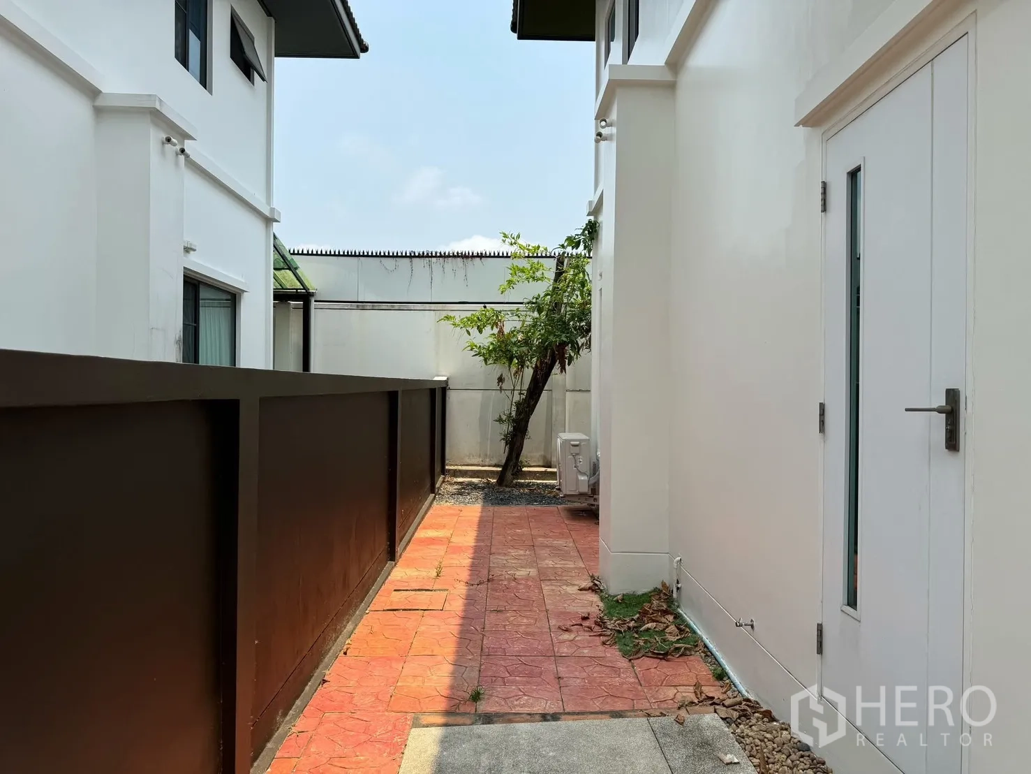 Detached House for rent in Prawet, Bangkok - Paved side yard with a white door, boundary walls and a small tree between houses.