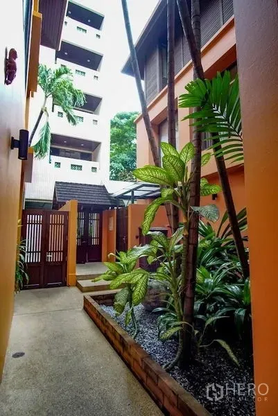 Townhouse for rent in Watthana, Bangkok - Shaded walkway beside lush garden leading to the townhouse’s wooden front gate.