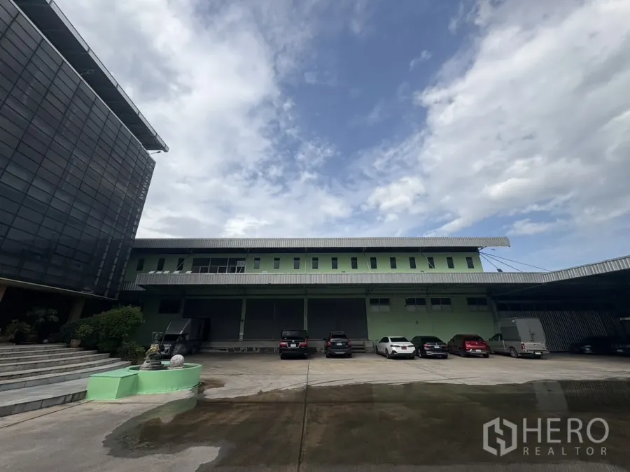 คลังสินค้า ให้เช่าในBang Khun Thian, Bangkok - ภาพด้านหน้าโกดังสีเขียว 2 ชั้น มีหลังคาคลุมท่าโหลด รถจอดหลายคัน และอาคารสำนักงาน ใต้ท้องฟ้ามีเมฆ