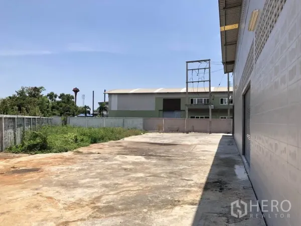 Factory for rent in Lat Lum Kaeo, Pathum Thani - Wide concrete yard beside the factory with boundary wall and greenery under clear sky