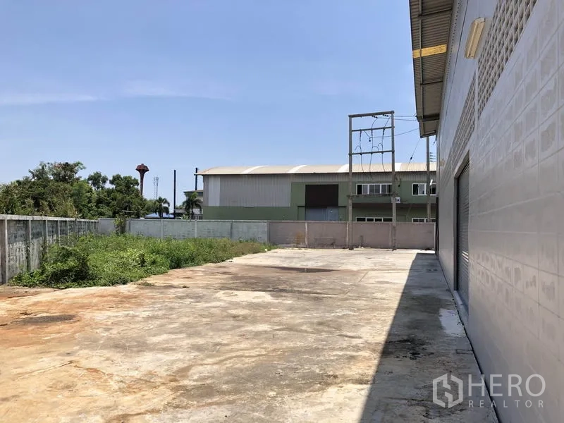 Factory for rent in Lat Lum Kaeo, Pathum Thani - Wide concrete yard beside the factory with boundary wall and greenery under clear sky
