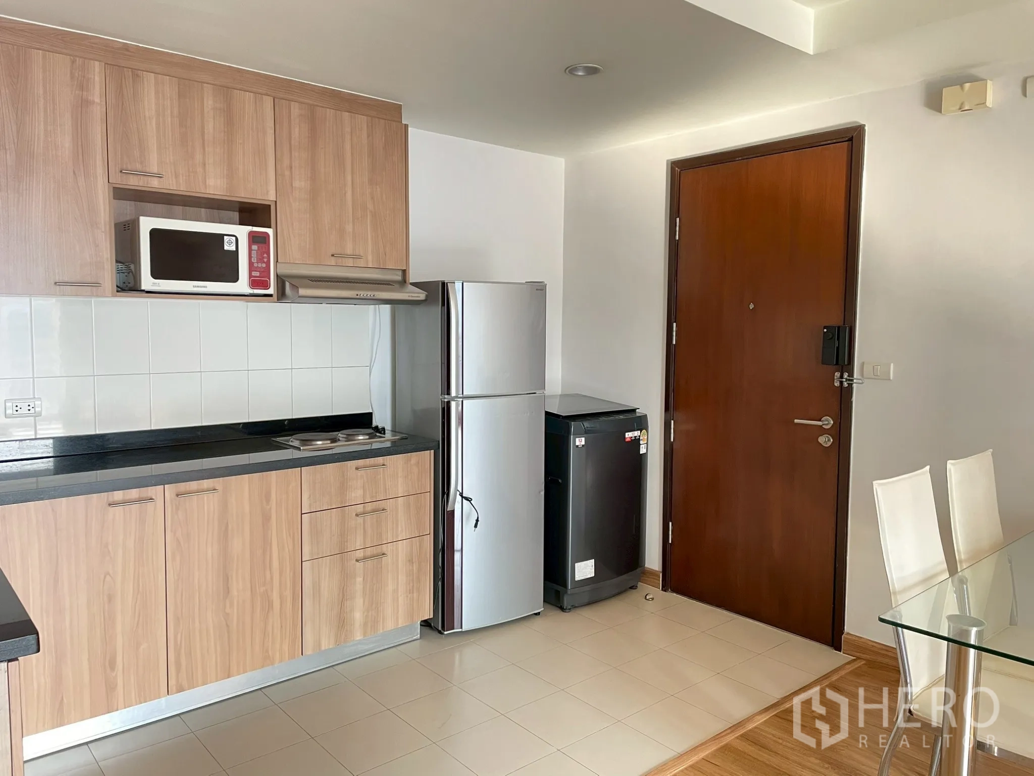 Apartment for rent in Khlong Toei, Bangkok - Kitchen detail showing cabinets, sink, cooktop, fridge, and entry door.