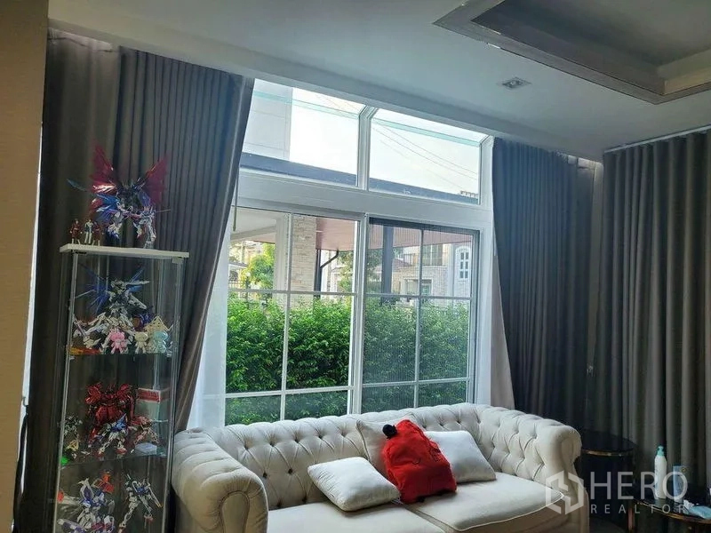 Detached House for sale in Bueng Kum, Bangkok - Living room with large windows, curtains and a tufted sofa facing greenery.