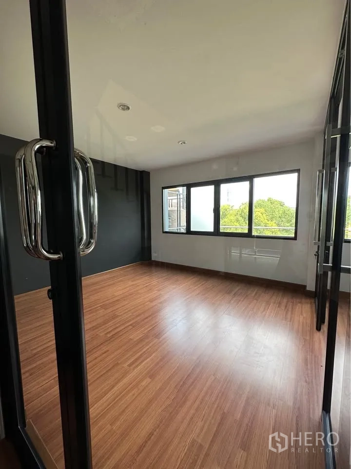Townhouse for rent in Pak Kret, Nonthaburi - Sunlit corner room with wood floors, black feature wall, and casement windows.