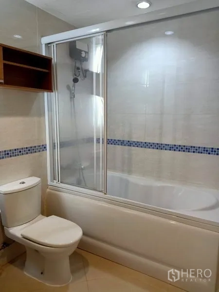 Condominium for rent in Bang Rak, Bangkok - Bathroom with bathtub, glass shower screen, toilet and wall shelf in neutral tiles.