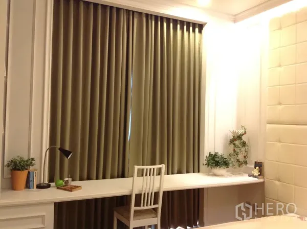 Condominium for rent in Watthana, Bangkok - Bedroom workspace with a long built-in desk, chair, potted plants, and full-length blackout curtains.