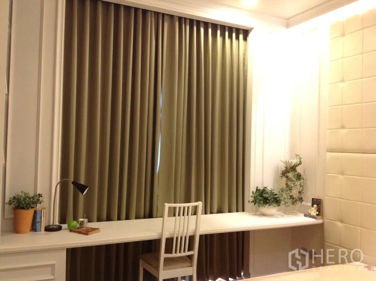Condominium for rent in Watthana, Bangkok - Bedroom workspace with a long built-in desk, chair, potted plants, and full-length blackout curtains.