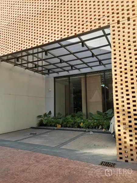 零售空间 出租在Khlong Toei, Bangkok - 现代零售建筑的有顶入口，玻璃店面与种植带，外立面为穿孔砖墙。