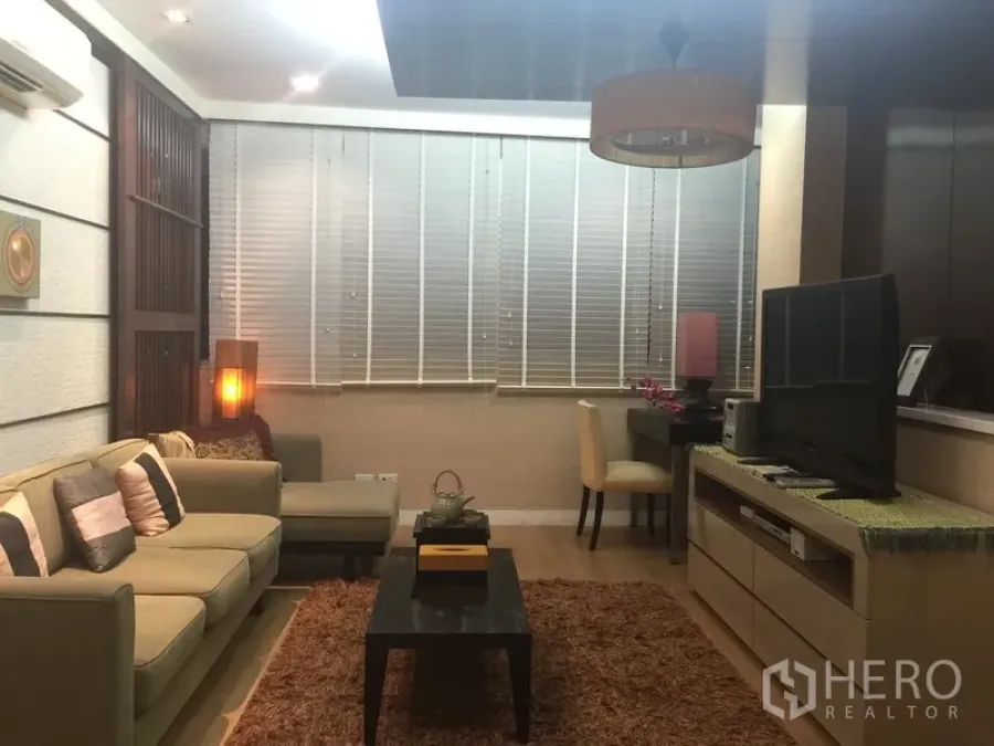 Condominium for rent or sale in Sathon, Bangkok - Modern living room with L-shaped sofa, TV console, desk and large window blinds.